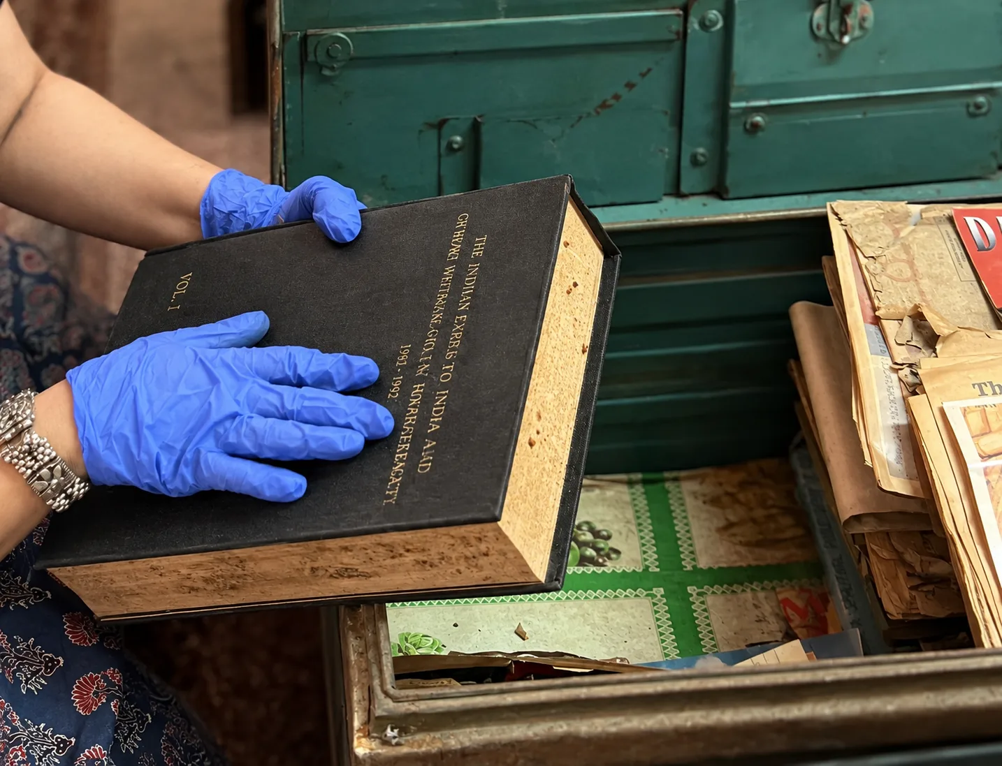 Preserving queer Indian archives Gloved hands lifting an old bound volume from a green metal trunk filled with papers and newsletters.